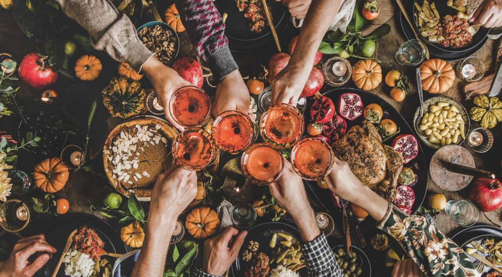 A table spread of fall produce and meals. Above the table six arms clink glasses full of orange liquid.